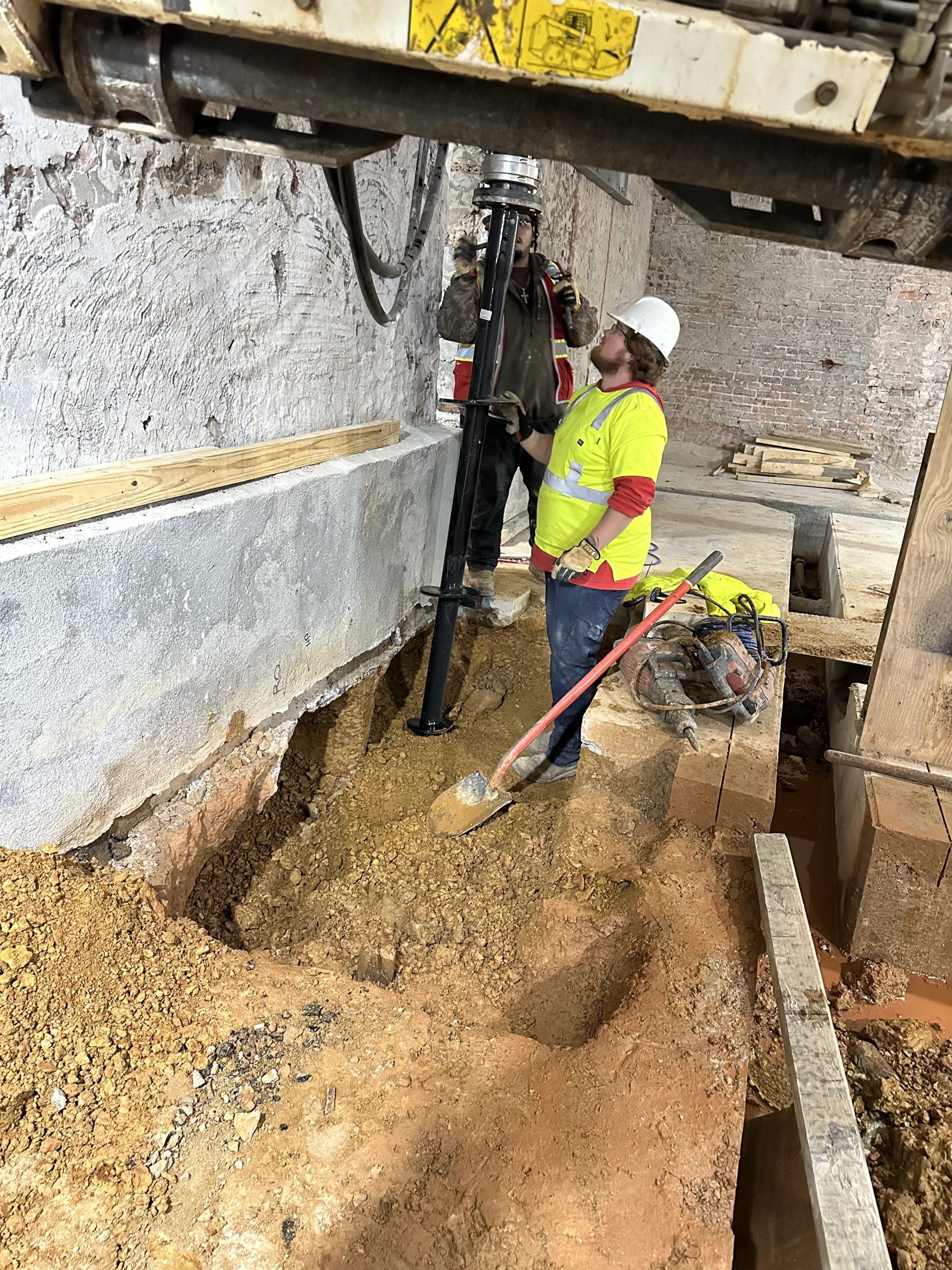 Helical pile being driven inside a basement excavation, worker looking up the shaft